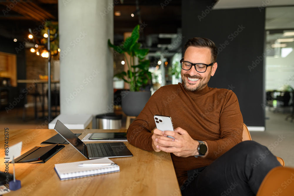 Young project manager reading e-mails over mobile phone while sitting by laptop and notepad at desk Young project manager reading e-mails over mobile phone while sitting by laptop and notepad at desk