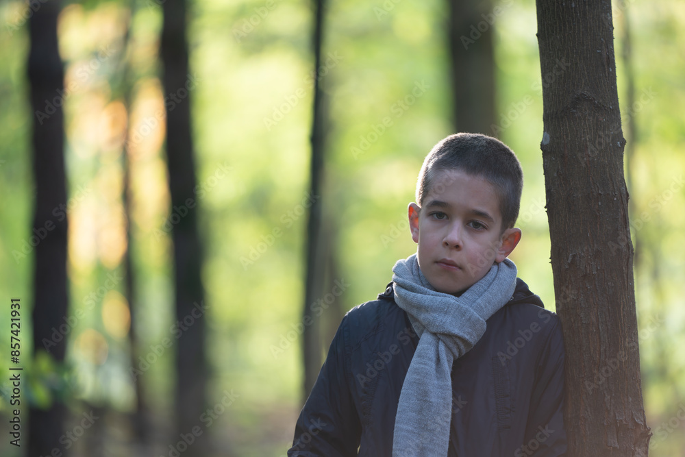 Young boy in dark jacket stands near tree in forest, looking at camera with neutral expression. Young boy in dark jacket stands near tree in forest, looking at camera with neutral expression.
