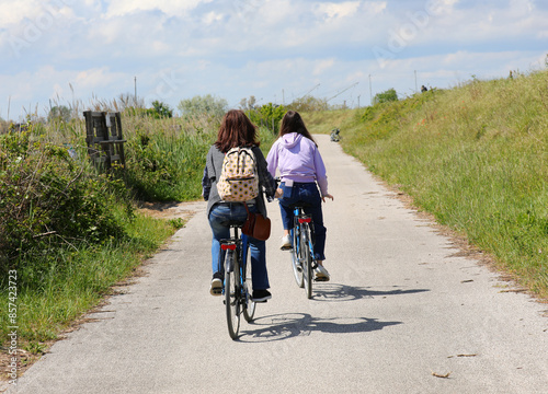 young mother woman and daughter pedaling on the cycle path in summer with a backpack  next to the river bank photo