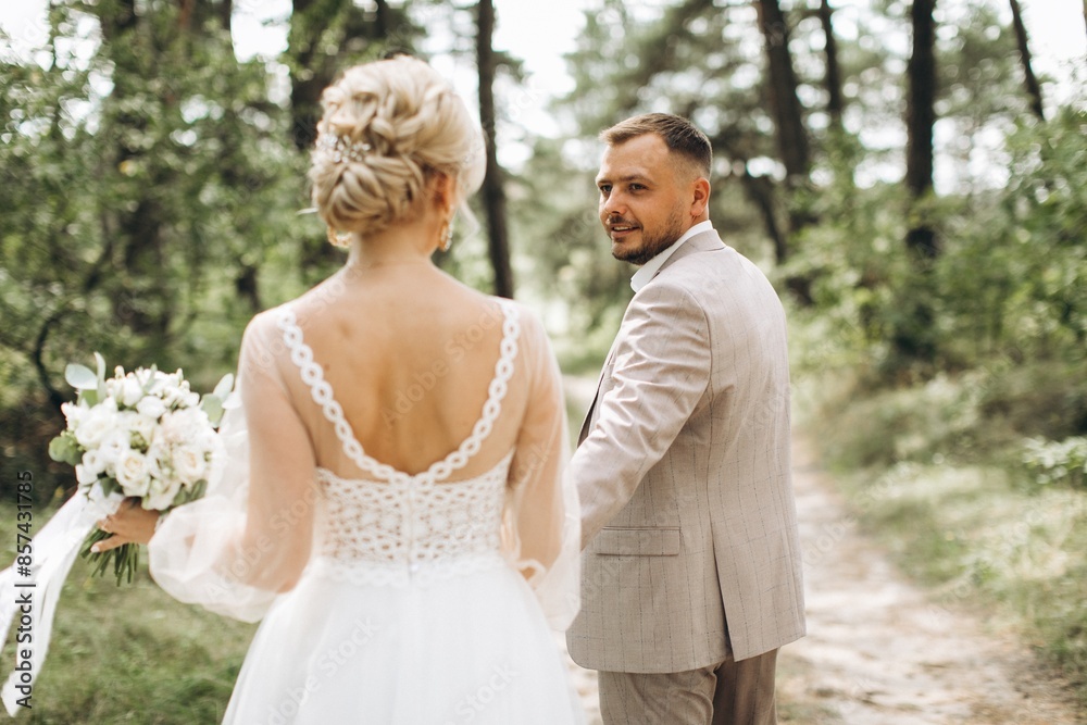 Fototapeta premium Wedding couple bride and groom walk hand in hand through a pine forest, sunlight filtering through the trees, highlighting their joyful expressions.