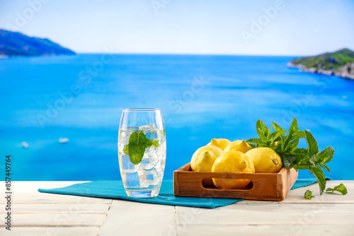 Lemon fruits, fresh mineral water, ice in glass on wooden table top with beautiful blur landscape of calm blue sea and blue sky. Vacation time and cool drink for hot day.