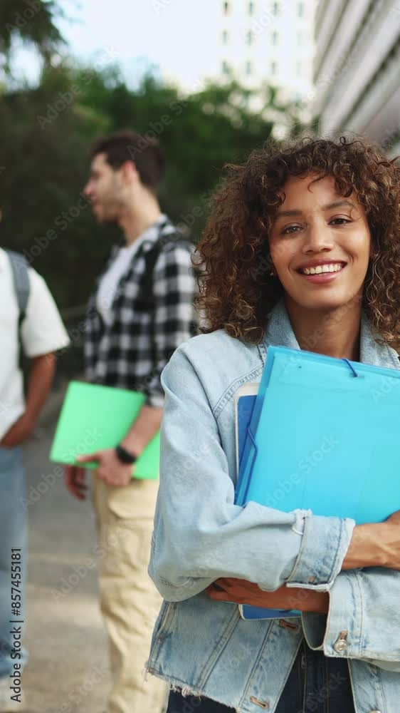 Portrait of a smiling young latin student looking at the camera while her classmates talk in the background on campus. Students socializing outdoors on campus, one smiling with folders Portrait of a smiling young latin student looking at the camera while her classmates talk in the background on campus. Students socializing outdoors on campus, one smiling with folders