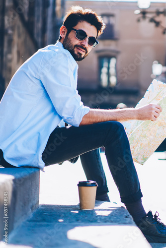 Portrait of cheerful bearded tourist in sunglasses sitting outdiirs on street of architectural city with old constructions while holding travel map for searching destination of showplaces photo