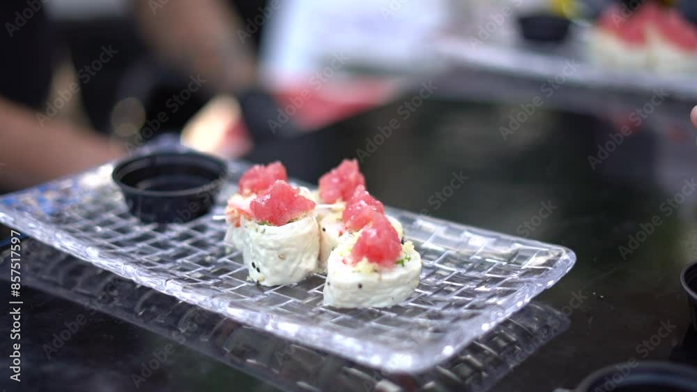 Un plato transparente con sushi de atún fresco y en el fondo las manos de un chef preparando mas comida japonesa