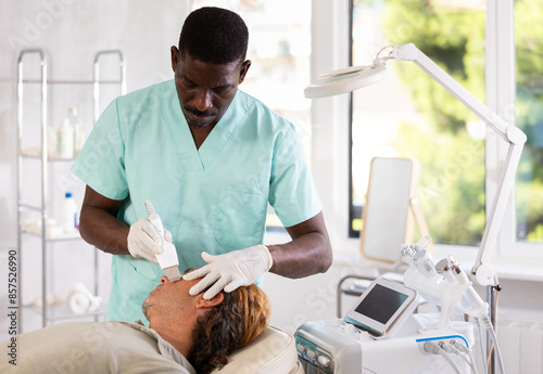 African American esthetician performing hardware cosmetic treatment for facial care on adult man in aesthetic medicine clinic, using ultrasonic shovel to cleanse skin. Professional male grooming photo