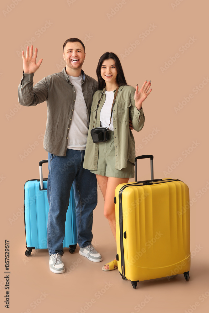 Beautiful young happy couple of tourists with suitcases waving hands on beige background Beautiful young happy couple of tourists with suitcases waving hands on beige background
