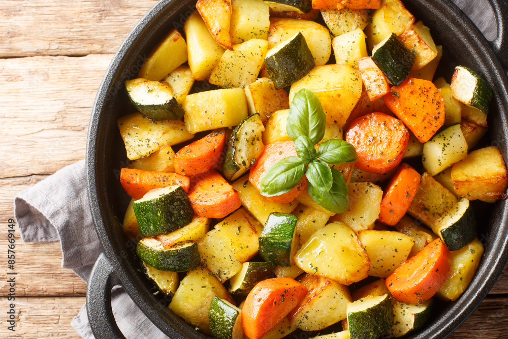 Vegetarian dish fried vegetables potatoes, carrots, zucchini and garlic close-up in a frying pan on a wooden table. Horizontal top view from above Vegetarian dish fried vegetables potatoes, carrots, zucchini and garlic close-up in a frying pan on a wooden table. Horizontal top view from above