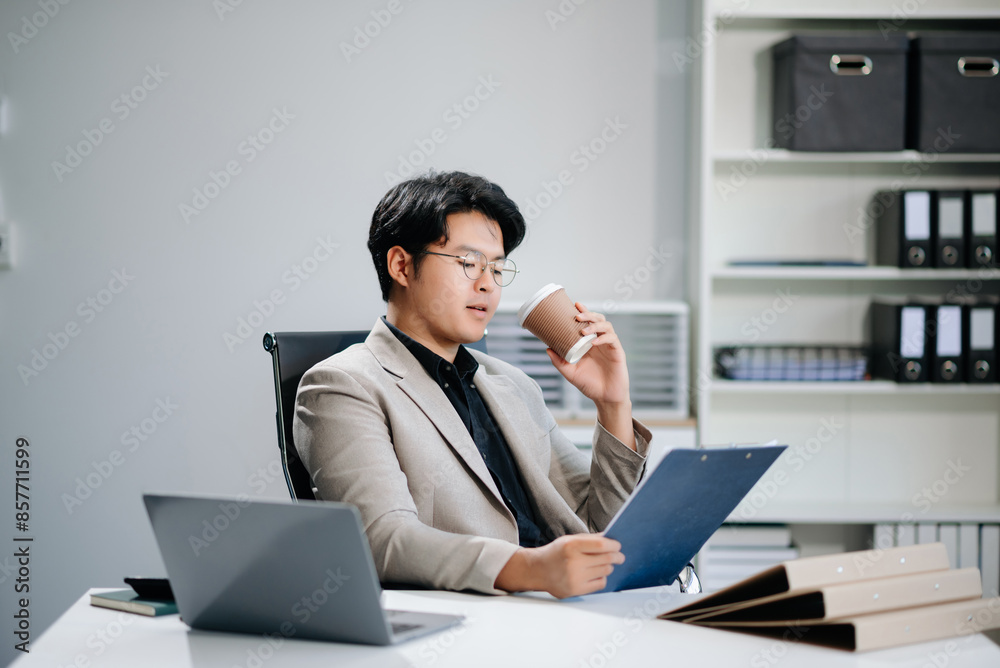 Confident Asian businessman typing laptop computer and digital tablet while holding coffee Confident Asian businessman typing laptop computer and digital tablet while holding coffee