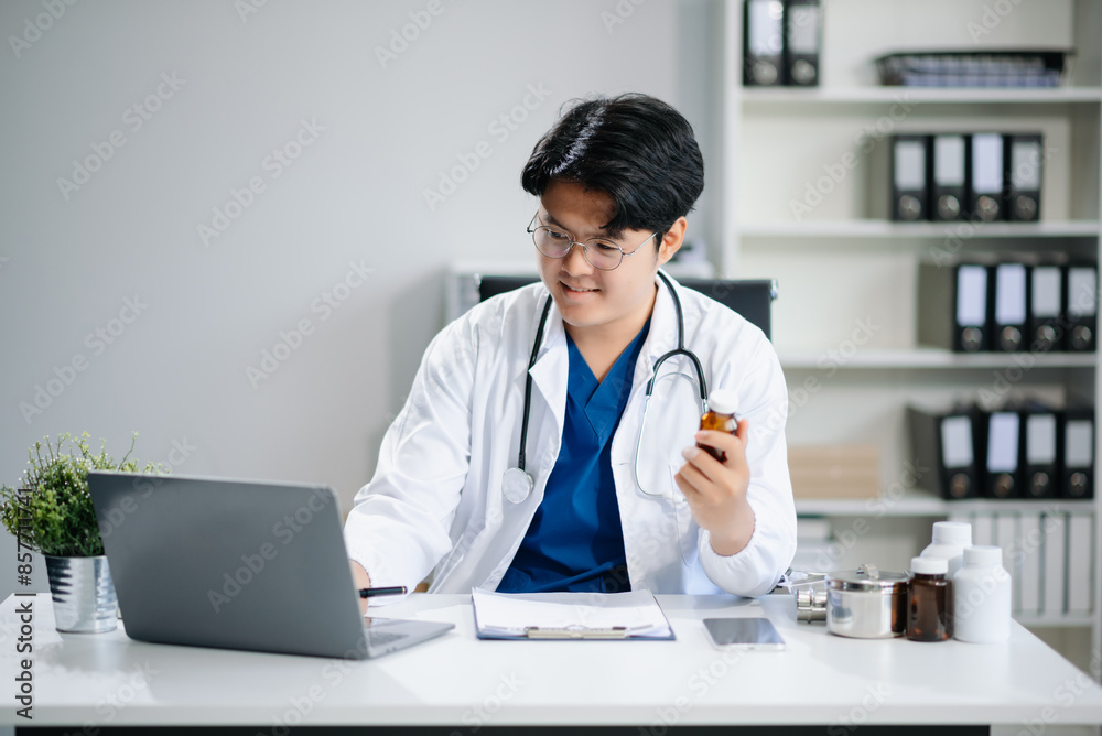 Confident young Asian male doctor in white medical uniform sit at desk working on computer. Smiling use laptop write Confident young Asian male doctor in white medical uniform sit at desk working on computer. Smiling use laptop write