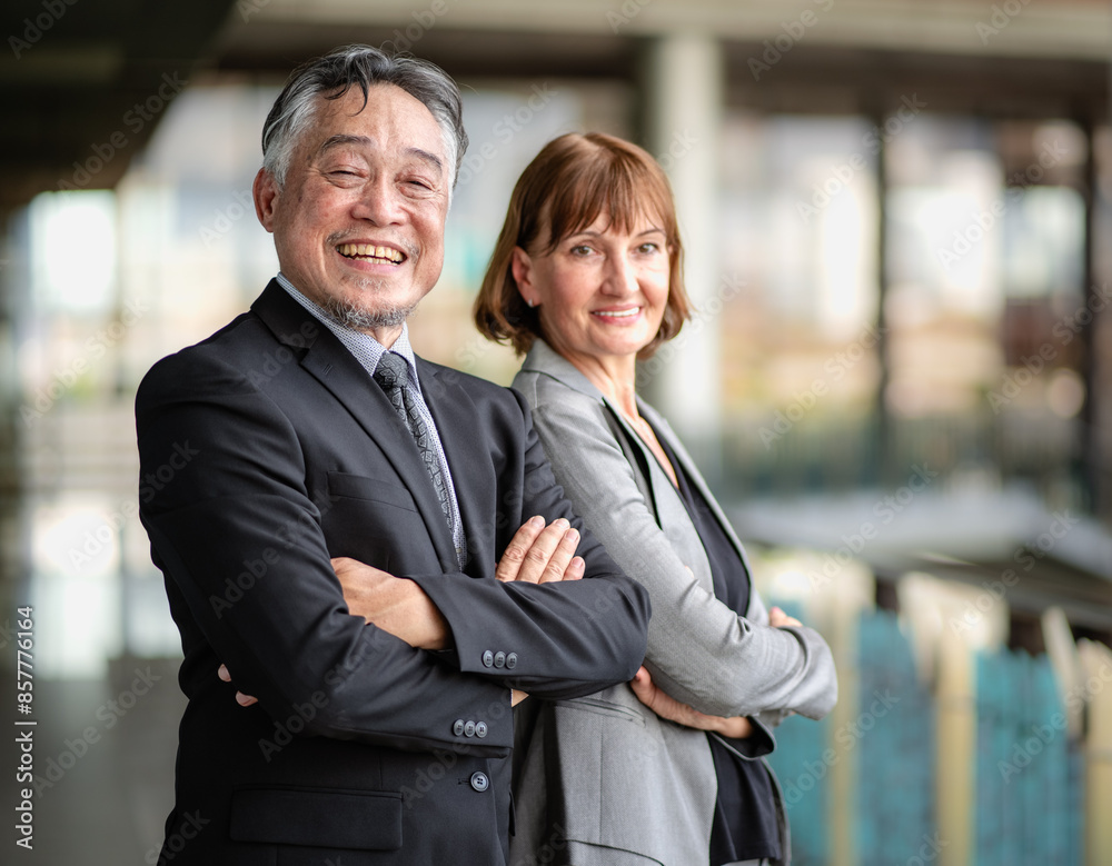Portrait of mature Asian businessman and Caucasian businesswoman standing outdoors arms crossed smiling. Diverse colleagues, well-dressed confident business person happy successful working together.