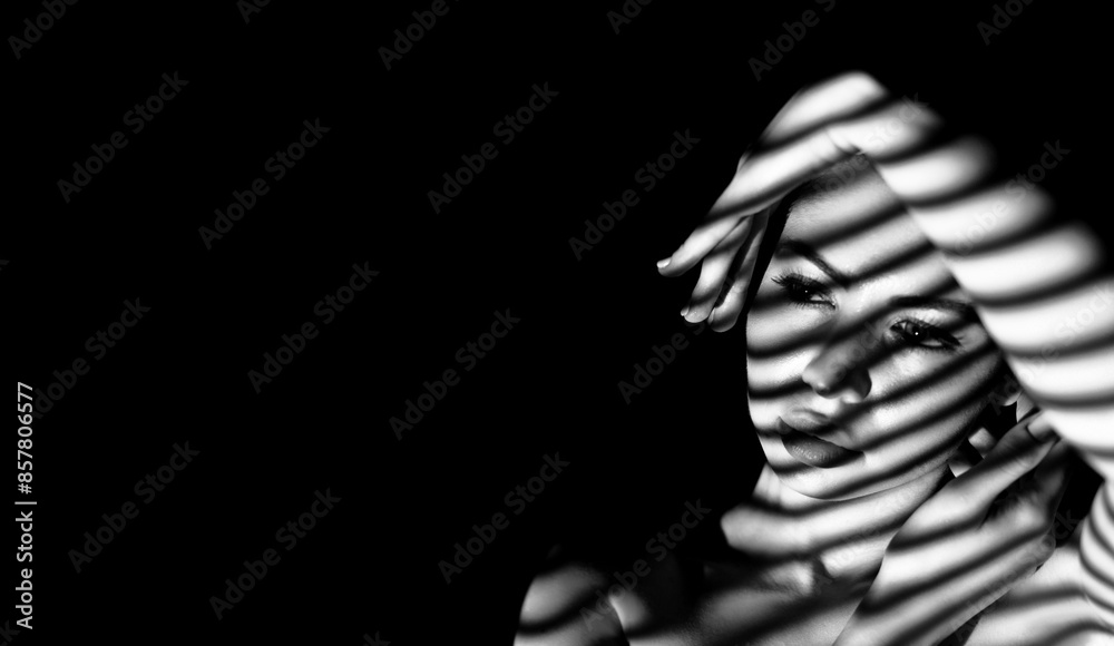 A black and white portrait of a woman in shadow. The light source casts striped shadows on her face and arm, creating a dramatic and artistic effect, copy space A black and white portrait of a woman in shadow. The light source casts striped shadows on her face and arm, creating a dramatic and artistic effect, copy space