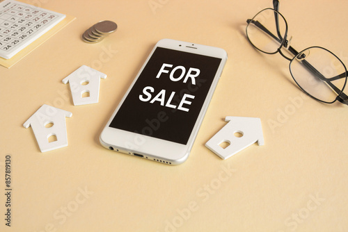 For sale symbol. symbol. Concept words FOR SALE written on the mobile phone screen. Beautiful beige background. Glasses, silver coins, miniature houses. Business, for sale concept, copy space.