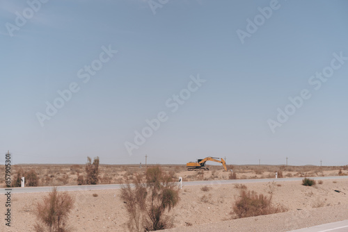 A large yellow excavator is on a dirt road in a desert. The sky is clear and the sun is shining brightly