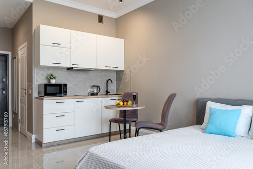 The interior of a small apartment with bed  blue pillows  light kitchen furniture  chairs with a table  kitchen appliances and refrigerator. apartment has gray walls.  fruits on the kitchen table.