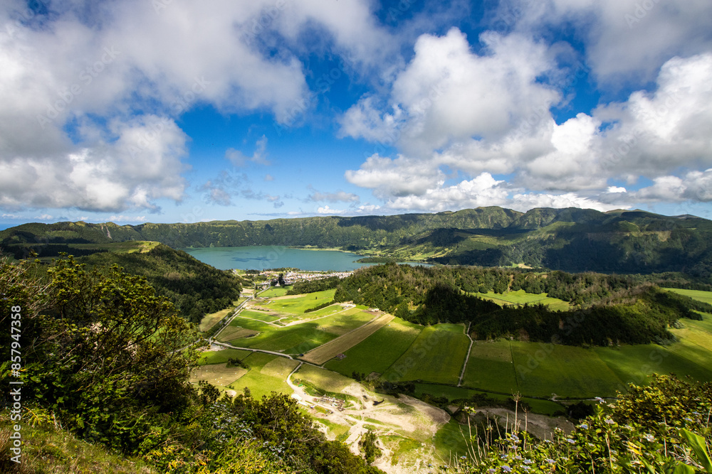 Obraz premium Aerial view of Sete Cidades in the Azores, showcasing a vibrant green valley, a blue crater lake, and lush surrounding mountains under a partly cloudy sky.