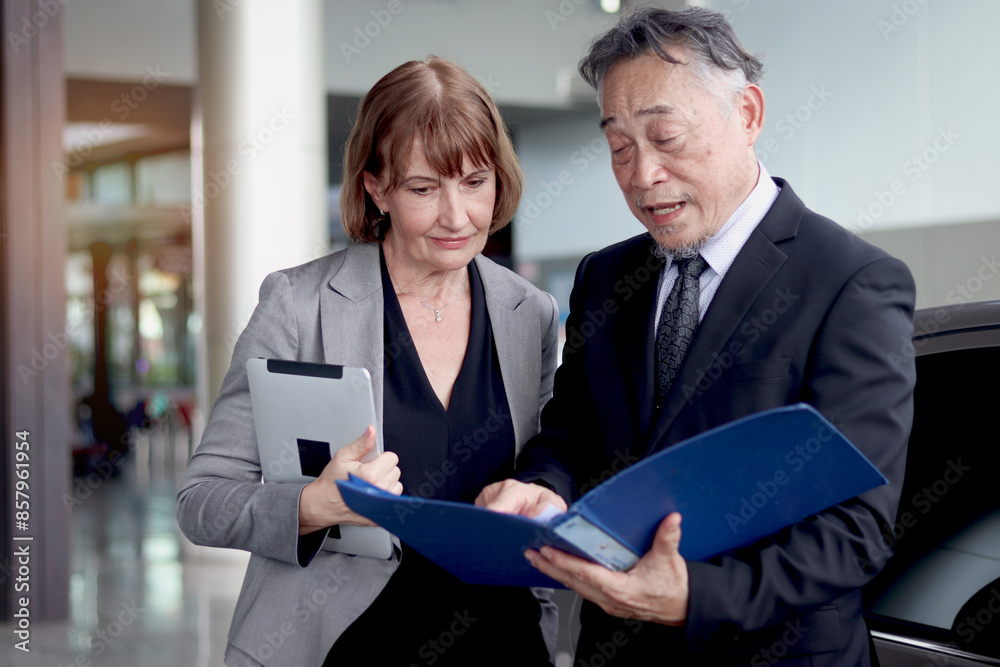 Asian senior businessman in suit holds document file to discuss information with his businesswoman partner. Elderly successful boss talks about schedule meeting with secretary at front office Asian senior businessman in suit holds document file to discuss information with his businesswoman partner. Elderly successful boss talks about schedule meeting with secretary at front office