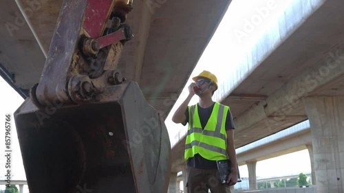 Asian male engineer standing next to an excavator Use the radio to check and order work at Construction size: highway bridge photo
