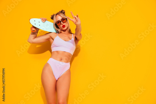 Young beautiful smiling woman posing near yellow wall in studio. Sexy model in swimwear bathing suit. Positive female with red lips. Holding penny skateboard. Happy and cheerful, shows peace sign