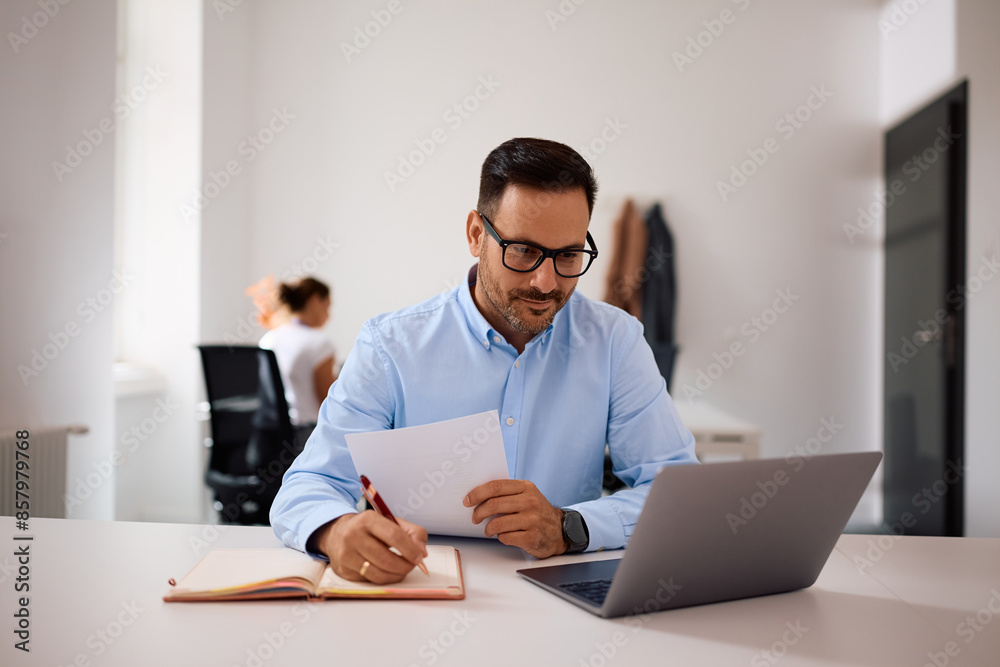 Smiling businessman writing notes while working on laptop in office. Smiling businessman writing notes while working on laptop in office.
