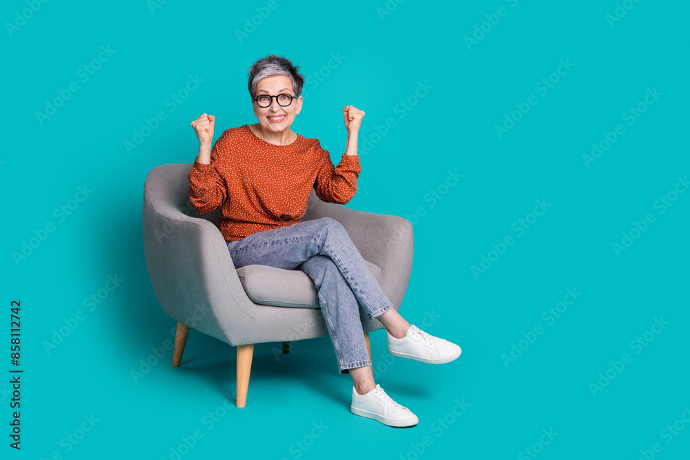 Full body photo of attractive senior woman sit armchair winning dressed stylish brown clothes isolated on cyan color background Full body photo of attractive senior woman sit armchair winning dressed stylish brown clothes isolated on cyan color background