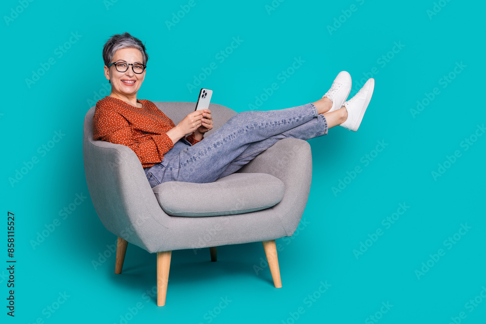 Full length photo of lovely pensioner lady sit armchair hold device dressed stylish brown garment isolated on cyan color background Full length photo of lovely pensioner lady sit armchair hold device dressed stylish brown garment isolated on cyan color background