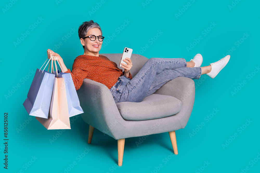 Full body photo of attractive senior woman sit armchair device shopper dressed stylish brown clothes isolated on cyan color background Full body photo of attractive senior woman sit armchair device shopper dressed stylish brown clothes isolated on cyan color background