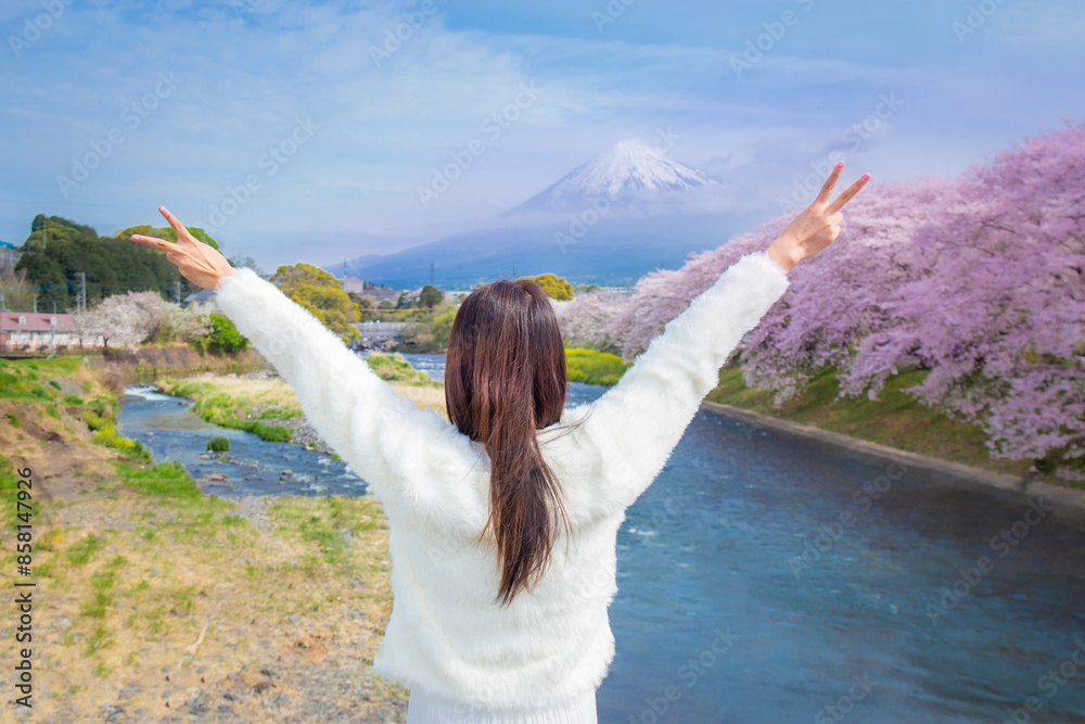 woman wearing a white dress stands with her arms outstretched and two fingers raised at a river with blooming cherry blossoms and Fuji in the foreground. woman wearing a white dress stands with her arms outstretched and two fingers raised at a river with blooming cherry blossoms and Fuji in the foreground.