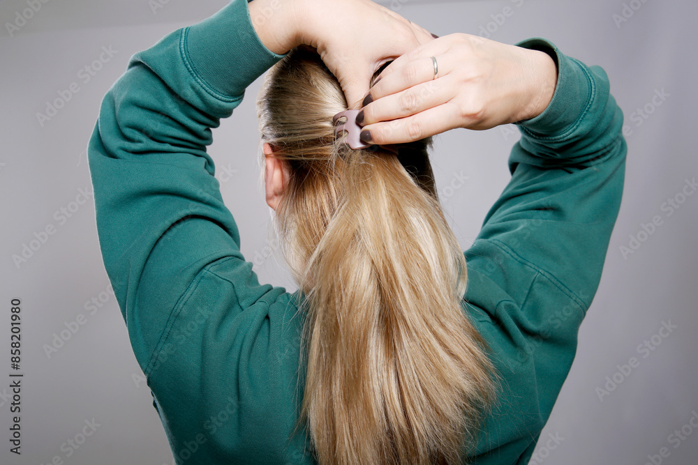 Fototapeta premium Close up back view of a woman with blond hair doing a hair claw clip in her hair, grey background