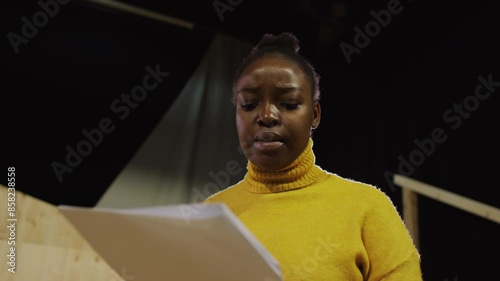 Low angle view of young talented Black actress reading monologue on paper and acting out with expression during rehearsal on theatre stage photo