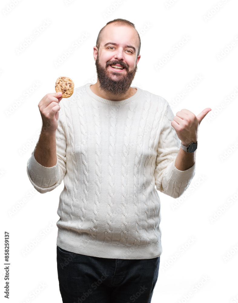 Young hipster man eating chocolate chips cookie over isolated background pointing and showing with thumb up to the side with happy face smiling Young hipster man eating chocolate chips cookie over isolated background pointing and showing with thumb up to the side with happy face smiling