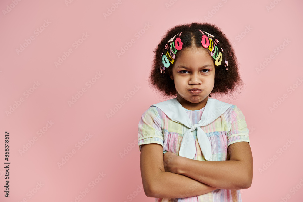 An African American girl with colorful hair clips stands with crossed arms in a pink and white dress on a pink background. An African American girl with colorful hair clips stands with crossed arms in a pink and white dress on a pink background.