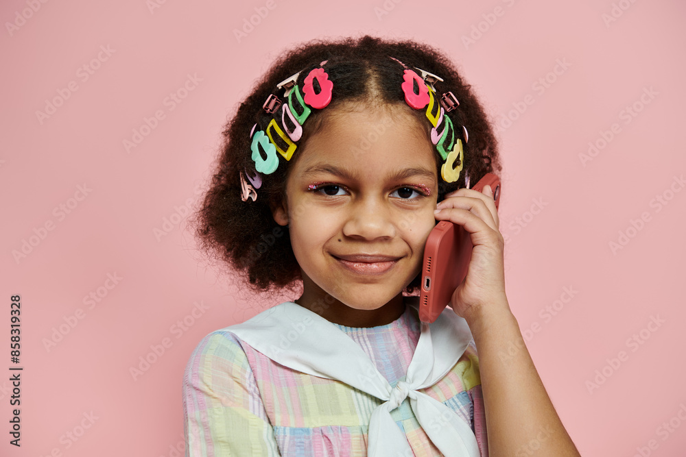 A young African American girl with colorful hair clips stands in a pink dress, smiling while talking on a pink phone. A young African American girl with colorful hair clips stands in a pink dress, smiling while talking on a pink phone.