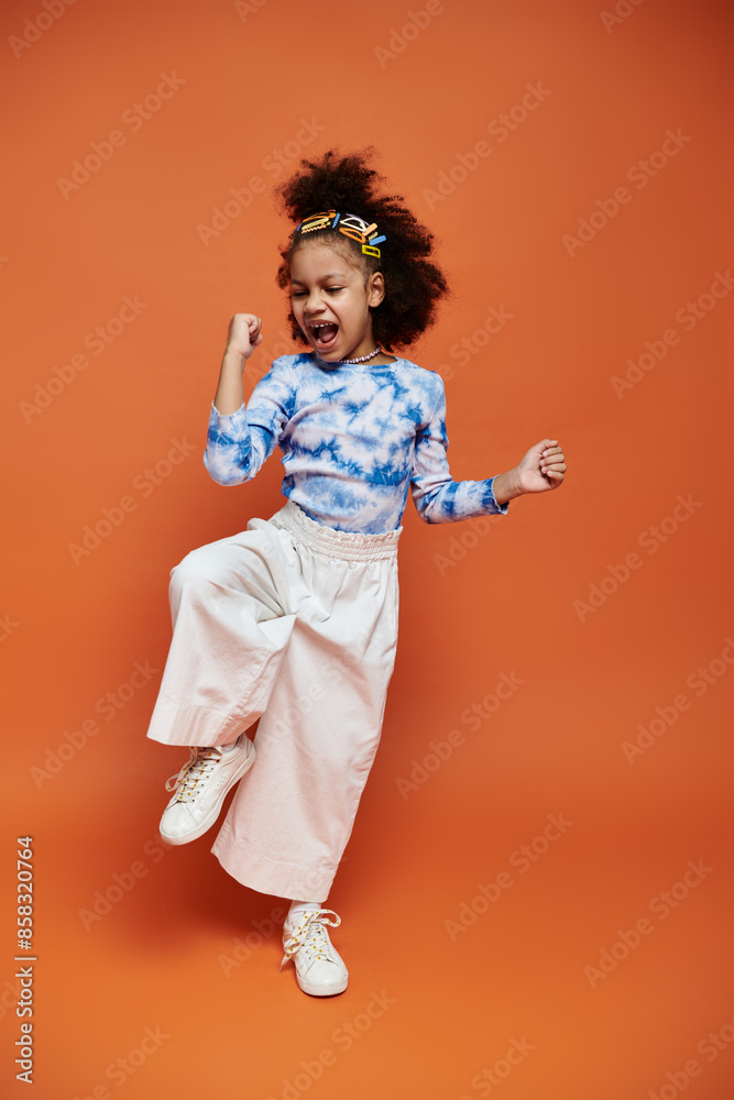 A young girl with stylish hair clips stands confidently in a trendy outfit, striking a joyful pose against an orange backdrop. A young girl with stylish hair clips stands confidently in a trendy outfit, striking a joyful pose against an orange backdrop.