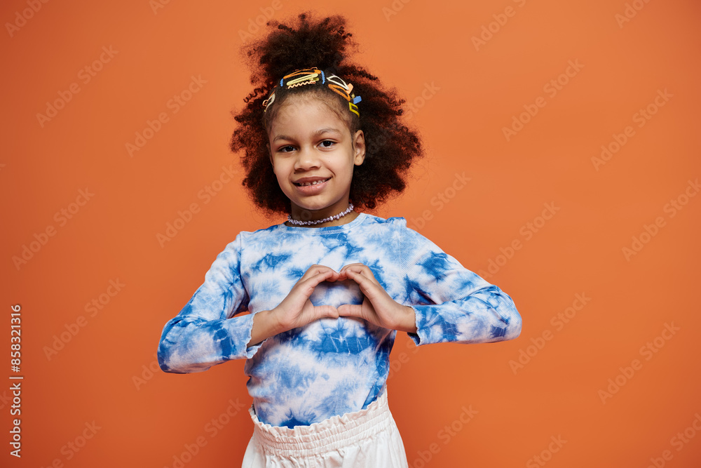 A young African American girl in a trendy outfit stands against a vibrant orange backdrop, smiling and making a heart shape with her hands. A young African American girl in a trendy outfit stands against a vibrant orange backdrop, smiling and making a heart shape with her hands.