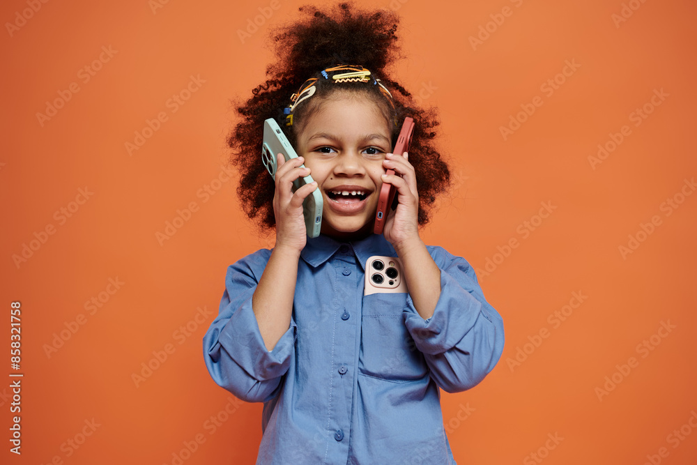 Obraz premium Young black girl with stylish hair clips, smiling playfully against an orange backdrop, holding two phones to her ears.
