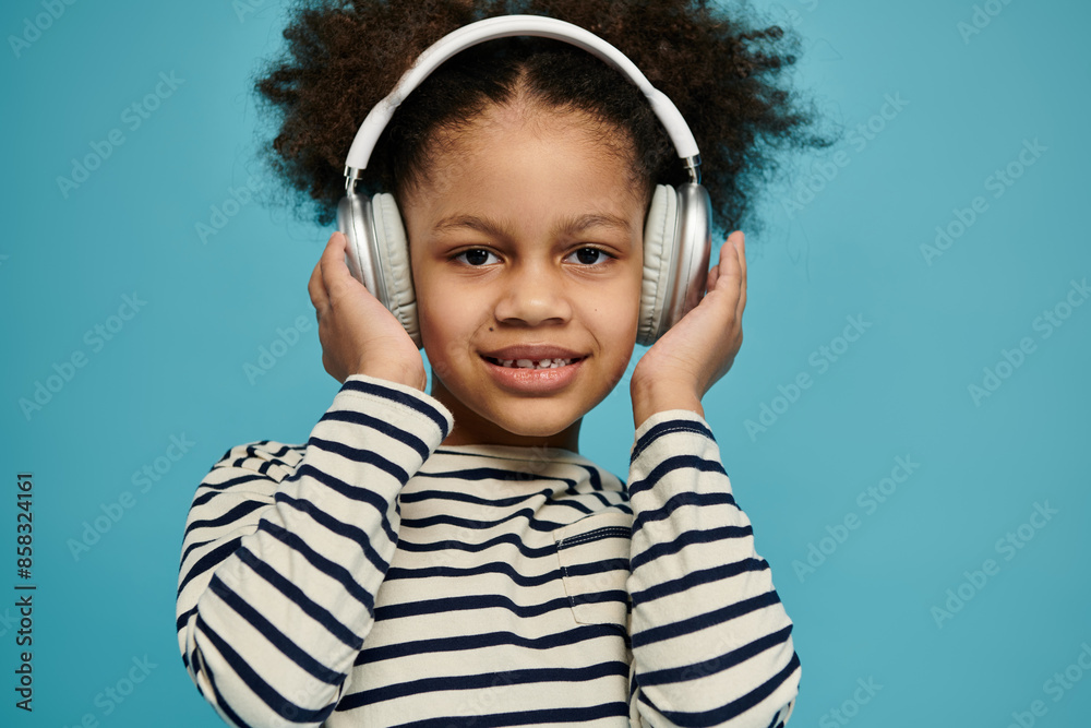 A smiling young Black girl with curly hair listens to music in headphones, wearing a striped shirt on a blue background. A smiling young Black girl with curly hair listens to music in headphones, wearing a striped shirt on a blue background.
