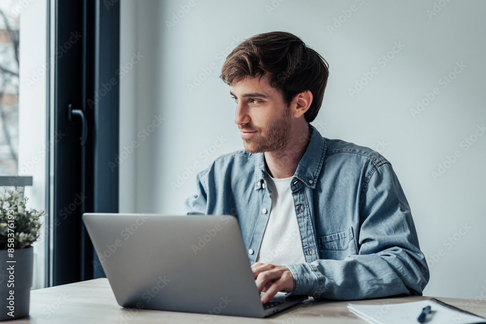 Side view handsome young businessman working with computer remotely, sitting at wooden table in office. Pleasant happy man communicating in social network, searching information online.. Side view handsome young businessman working with computer remotely, sitting at wooden table in office. Pleasant happy man communicating in social network, searching information online..
