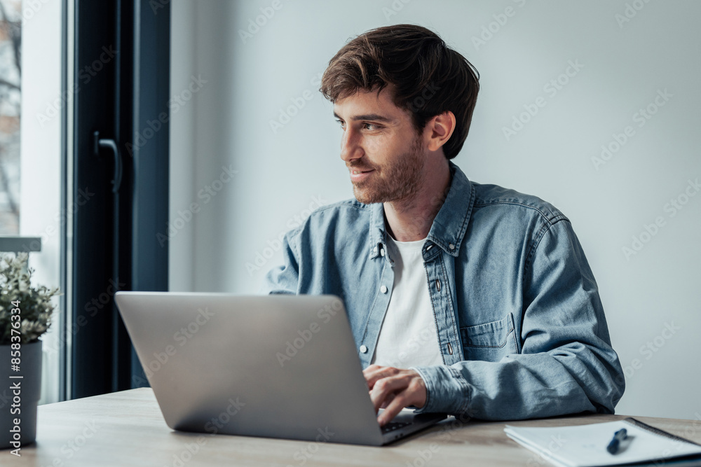 Side view handsome young businessman working with computer remotely, sitting at wooden table in office. Pleasant happy man communicating in social network, searching information online.. Side view handsome young businessman working with computer remotely, sitting at wooden table in office. Pleasant happy man communicating in social network, searching information online..