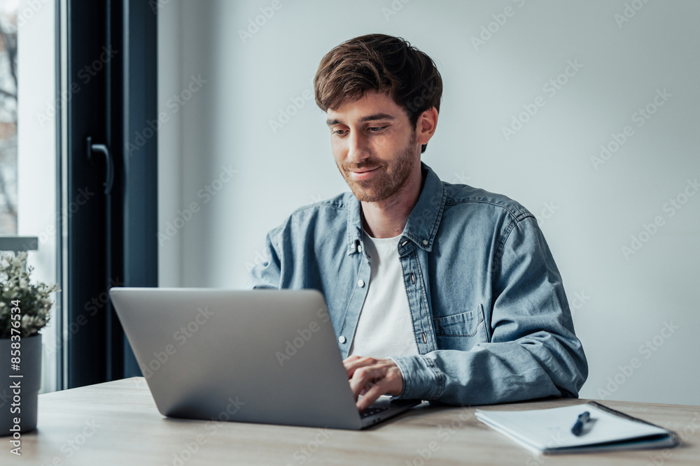 Side view handsome young businessman working with computer remotely, sitting at wooden table in office. Pleasant happy man communicating in social network, searching information online.. Side view handsome young businessman working with computer remotely, sitting at wooden table in office. Pleasant happy man communicating in social network, searching information online..