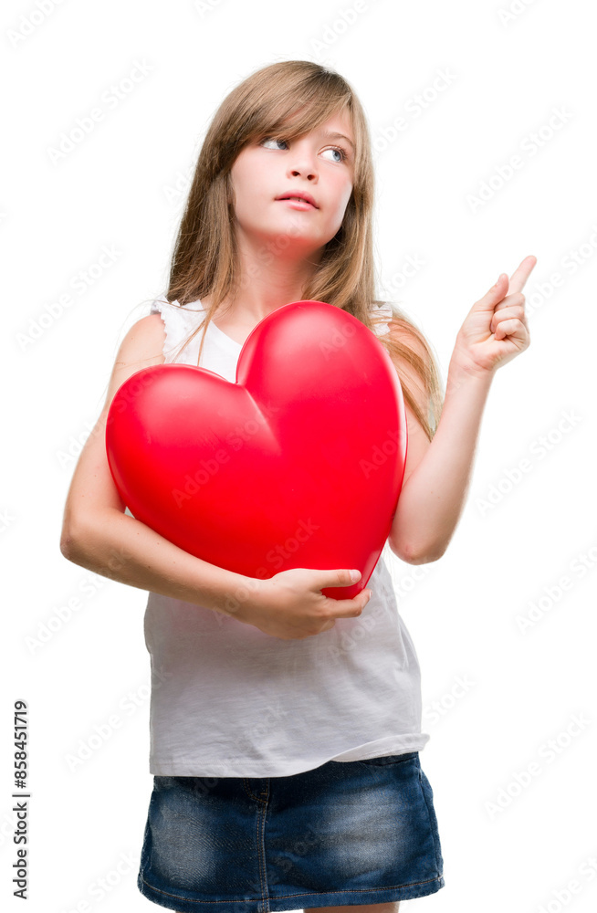 Young blonde toddler holding a red heart very happy pointing with hand and finger to the side Young blonde toddler holding a red heart very happy pointing with hand and finger to the side