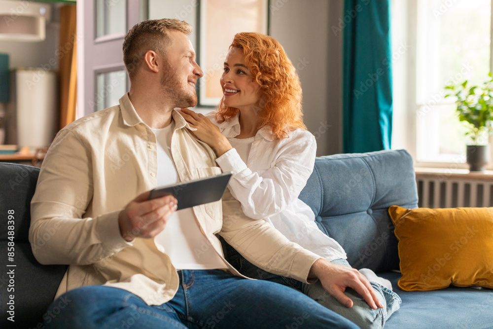 Happy young couple relaxing on couch in living room and using tablet gadget for shopping online, looking at each other and chatting Happy young couple relaxing on couch in living room and using tablet gadget for shopping online, looking at each other and chatting