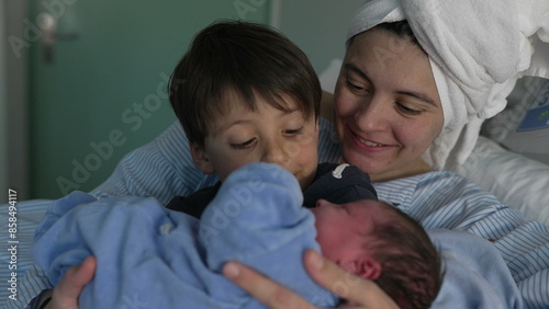 Mother and child welcoming newborn baby in arms at hospital after childbirth. nurse handing little infant to brother's arm next to mom at clinic, siblings bonding for the first time