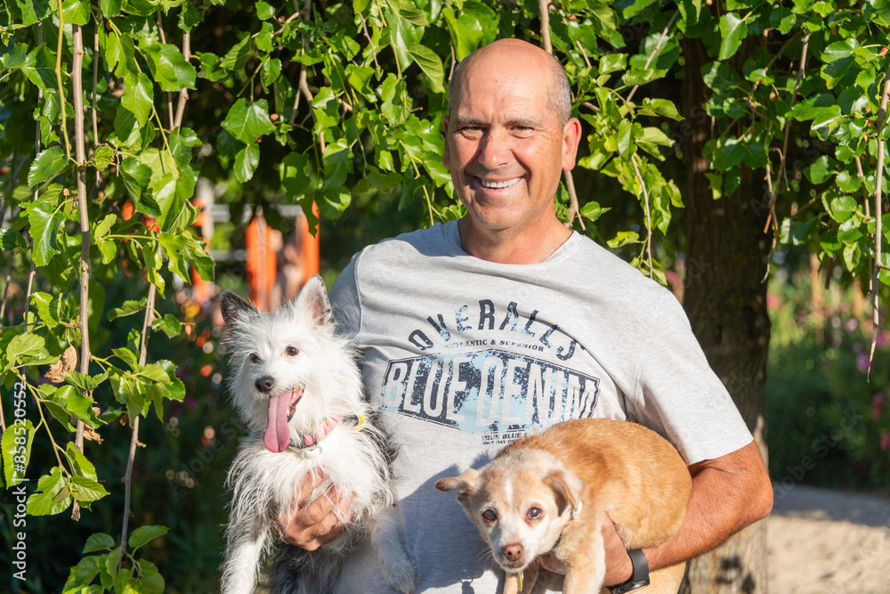 Obraz premium Cheerful man holding two small dogs, one with fluffy black and white fur and the other with light brown fur, in a sunlit garden.