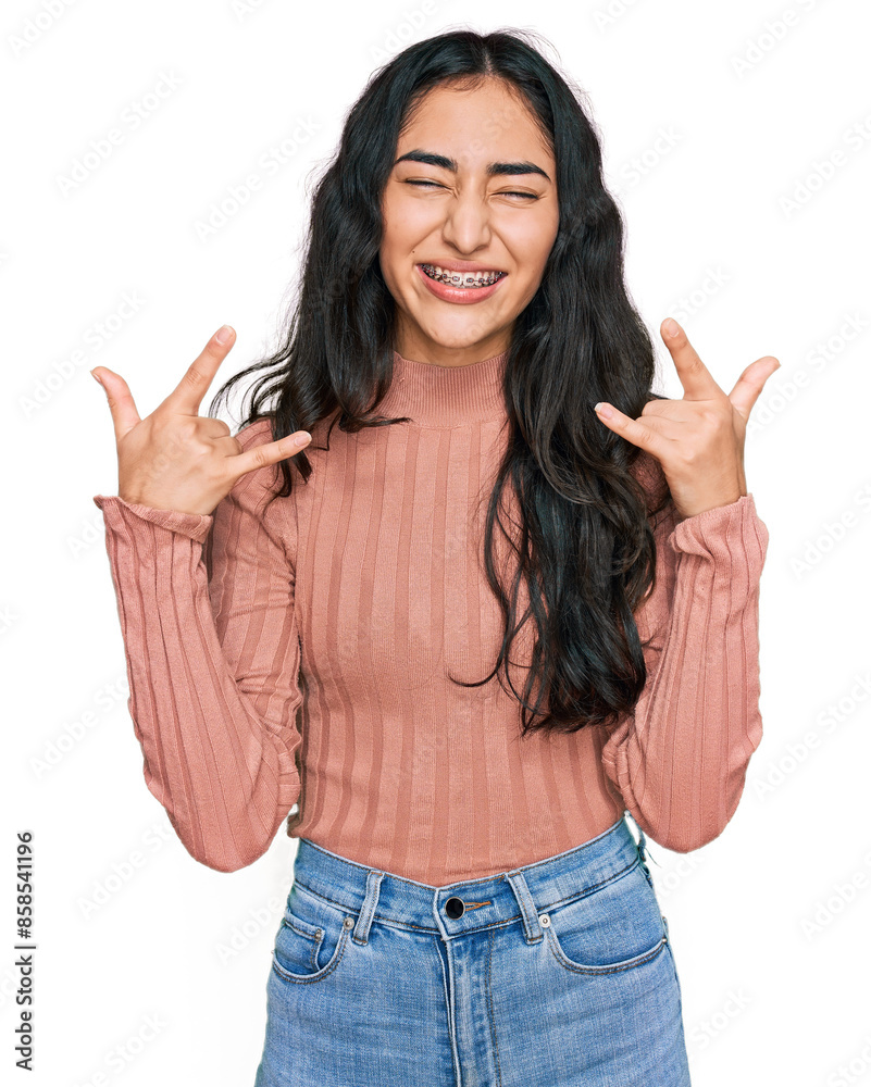 Hispanic teenager girl with dental braces wearing casual clothes shouting with crazy expression doing rock symbol with hands up. music star. heavy concept. Hispanic teenager girl with dental braces wearing casual clothes shouting with crazy expression doing rock symbol with hands up. music star. heavy concept.