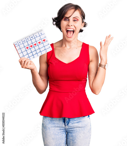 Beautiful young woman with short hair holding heart calendar celebrating victory with happy smile and winner expression with raised hands photo