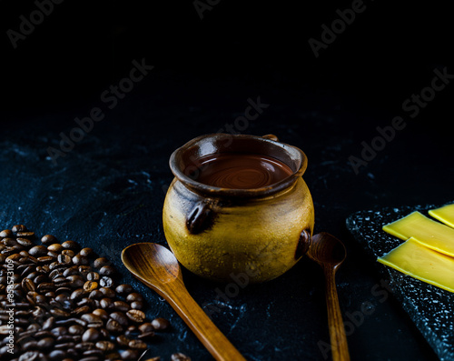 cup of chocolate in a yellow ceramic mug on a black table with coffee beans on the left side and a tray with slices of yellow cheese on the right
