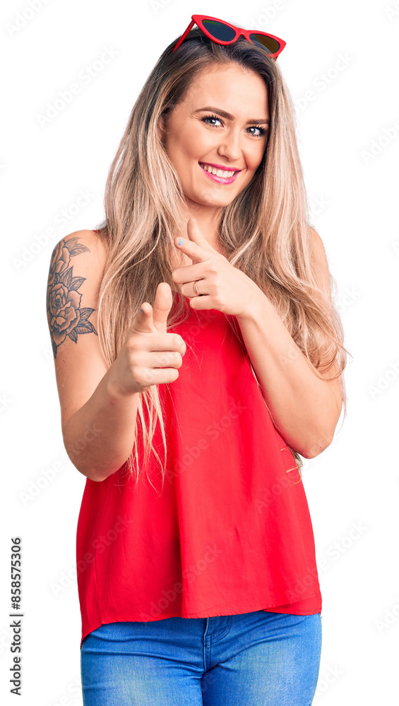 Young beautiful blonde woman wearing sleeveless t-shirt and sunglasses pointing fingers to camera with happy and funny face. good energy and vibes. Young beautiful blonde woman wearing sleeveless t-shirt and sunglasses pointing fingers to camera with happy and funny face. good energy and vibes.
