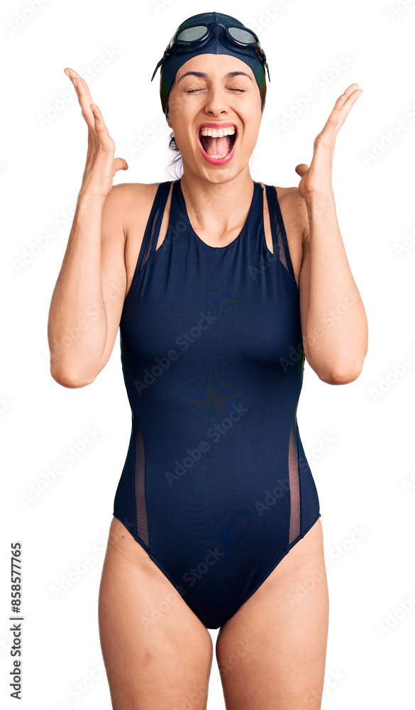 Young beautiful hispanic woman wearing swimwear and swimmer glasses celebrating mad and crazy for success with arms raised and closed eyes screaming excited. winner concept
