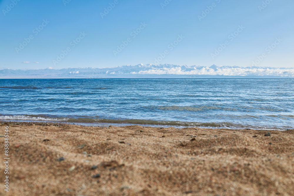 Sandy beach, coast of southern shore of big lake Issyk-Kul, summer vacation in Kyrgyzstan. Clear blue sky, emerald colored water of pond, small wave. Kirgiziya travel destination. Mountains in clouds Sandy beach, coast of southern shore of big lake Issyk-Kul, summer vacation in Kyrgyzstan. Clear blue sky, emerald colored water of pond, small wave. Kirgiziya travel destination. Mountains in clouds