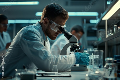 A scientist in a lab coat and safety glasses closely examines samples through a microscope in a modern laboratory.